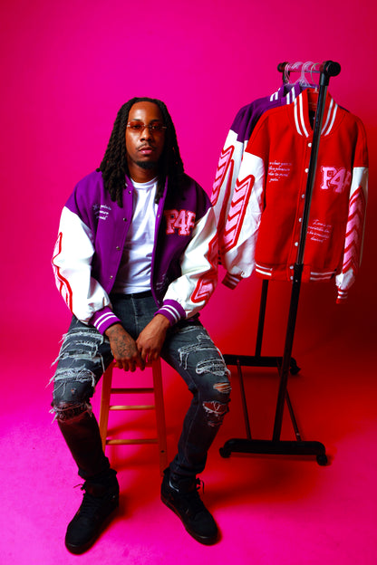 Person wearing a purple varsity jacket sitting next to a display of similar jackets on a pink background