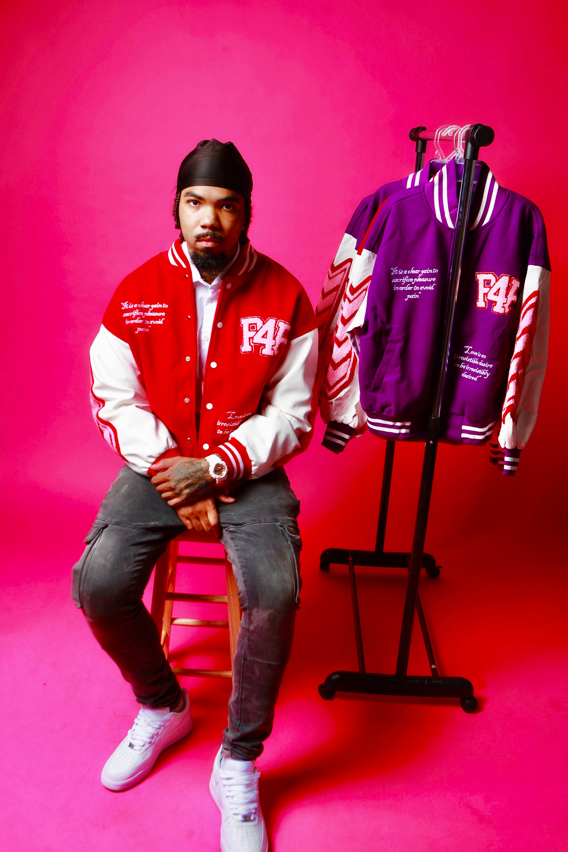 Man sitting on a stool next to a clothing rack wearing a red and white jacket with 'P4P' branding against a pink background.