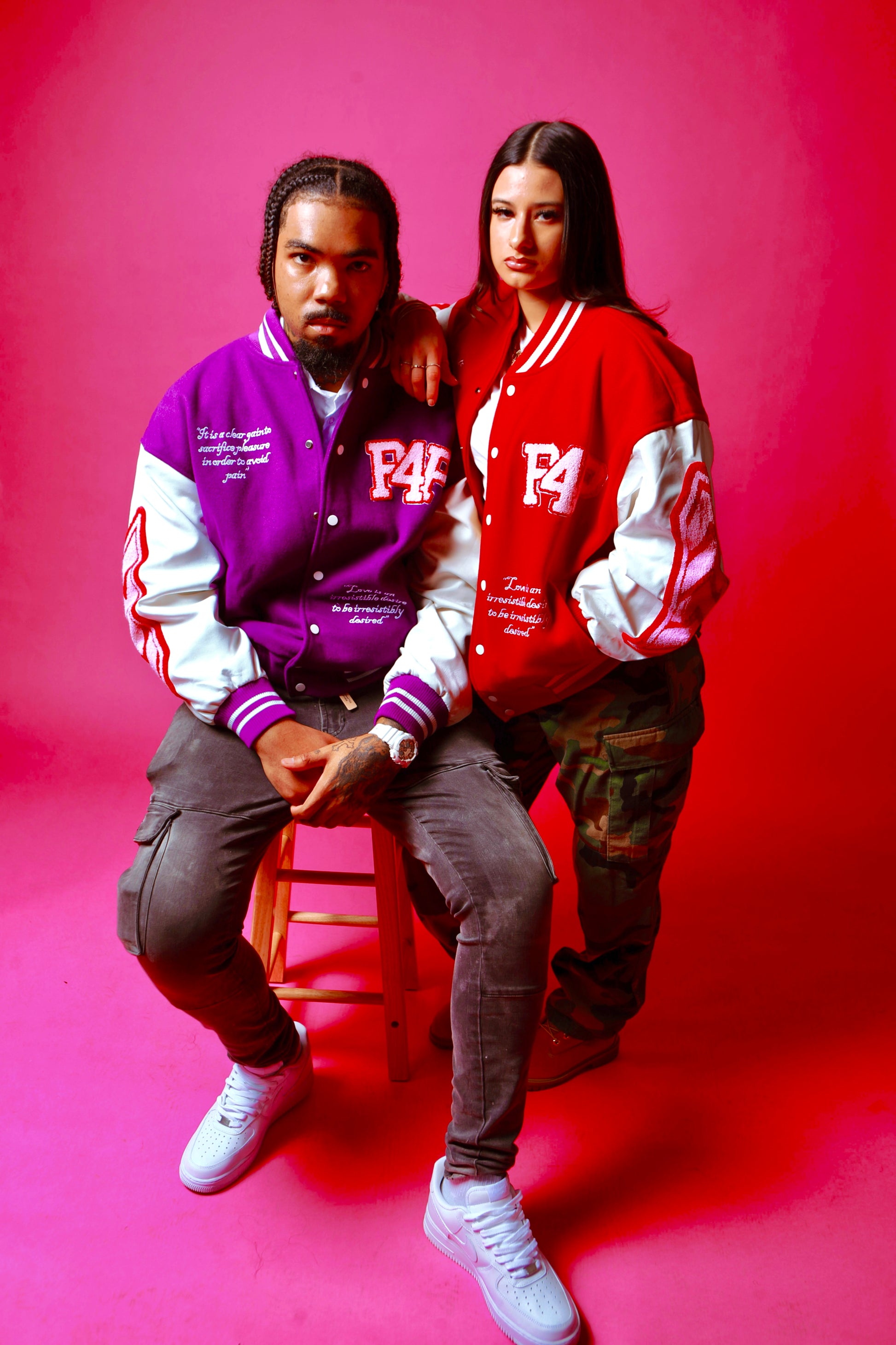Two people wearing colorful red, purple and pink streetwear letterman jackets sitting on a stool against a pink background